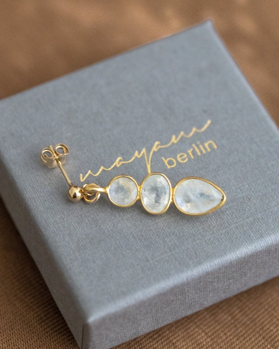 Ein goldener Ohrstecker mit einem dreifachen Mondstein-Anhänger liegt auf einem grauen Schmucketui.