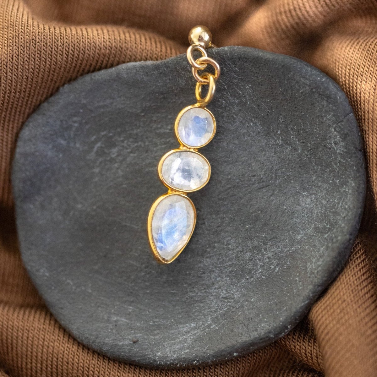 Ein goldener Ohrstecker mit einem dreifachen Mondstein-Anhänger lehnt an einem grauen Steinschälchen, das auf braunem Stoff liegt.