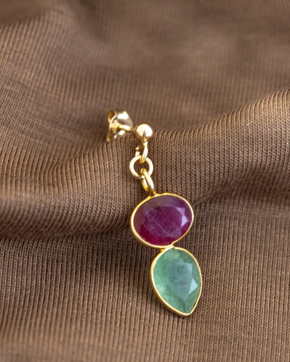 Ein Ohrstecker, der aus einem gold gerahmten Rubin und einem darunter angebrachten, ebenfalls gold gefassten, Prehnit besteht, lehnt an braunem Stoff.