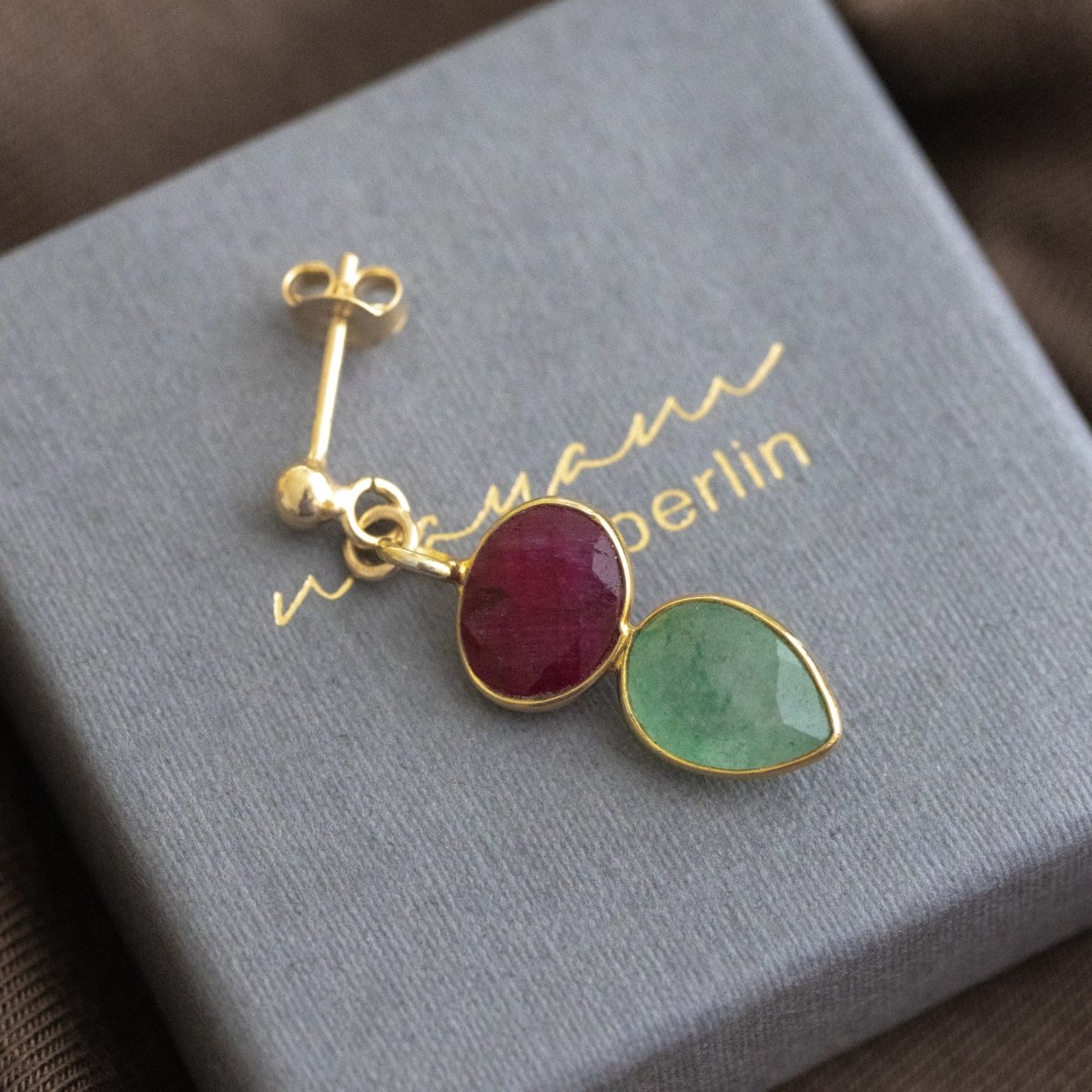 Ein Ohrstecker, der aus einem gold gerahmten Rubin und einem darunter angebrachten, ebenfalls gold gefassten, Prehnit besteht, liegt auf einem grauen Schmucketui.