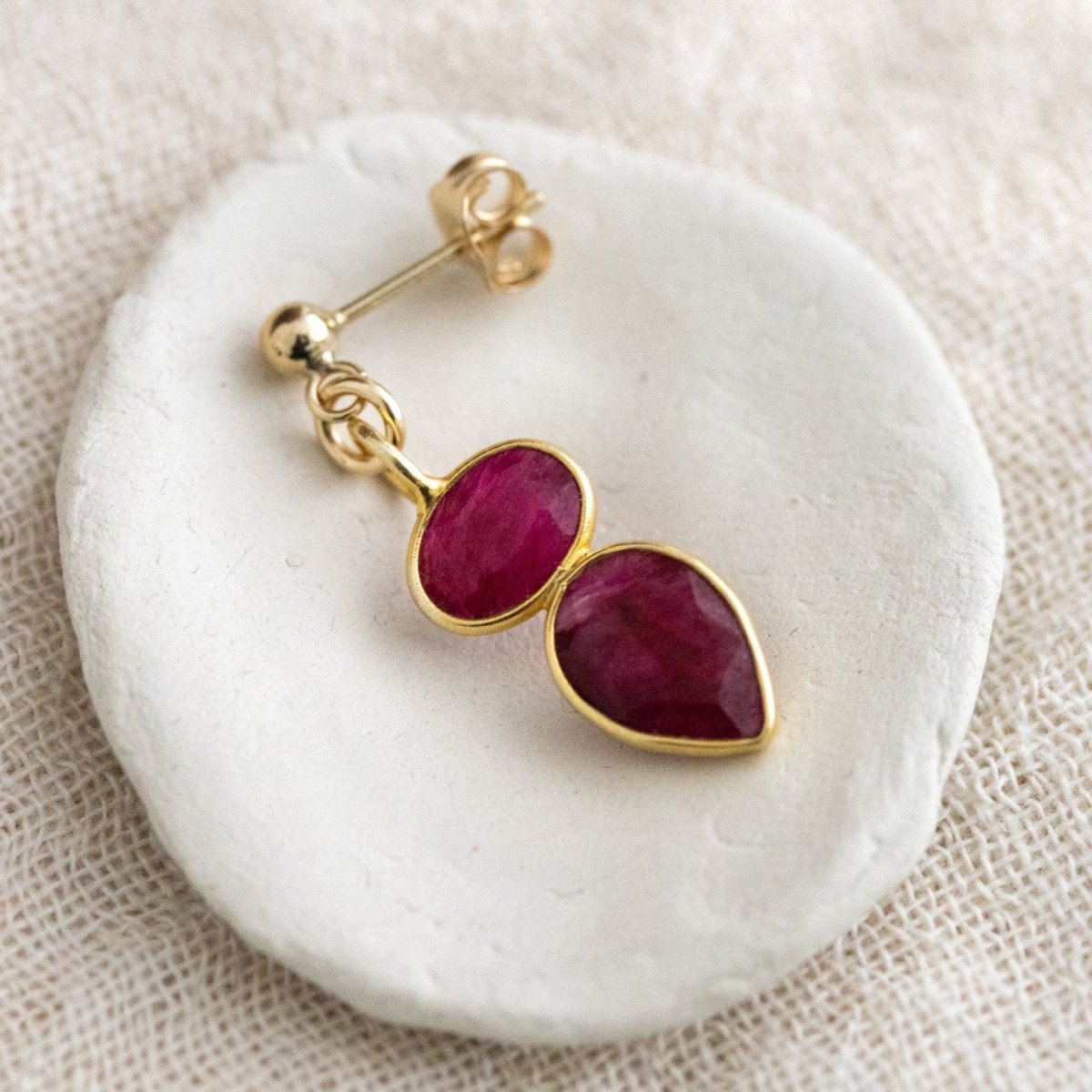 Ein goldener Ohrstecker mit zwei übereinander angebrachten Rubinen liegt in einem weißen Schälchen auf cremefarbenen Leinenstoff.