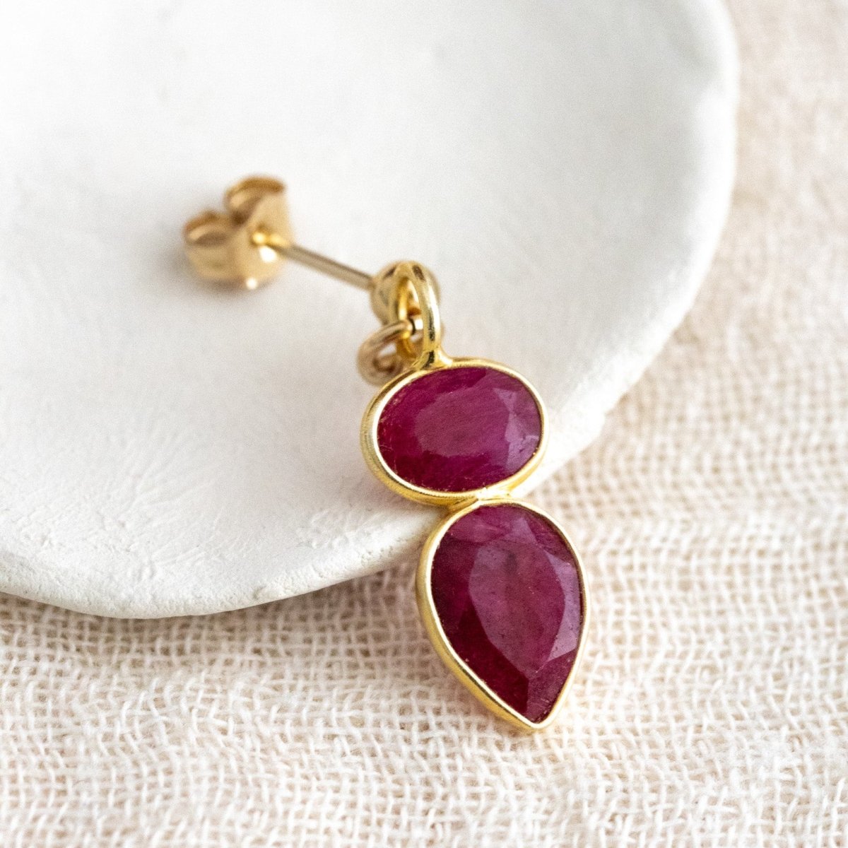 Ein goldener Ohrstecker mit zwei übereinander angebrachten Rubinen lehnt an einem weißen Schälchen auf cremefarbenen Leinenstoff.