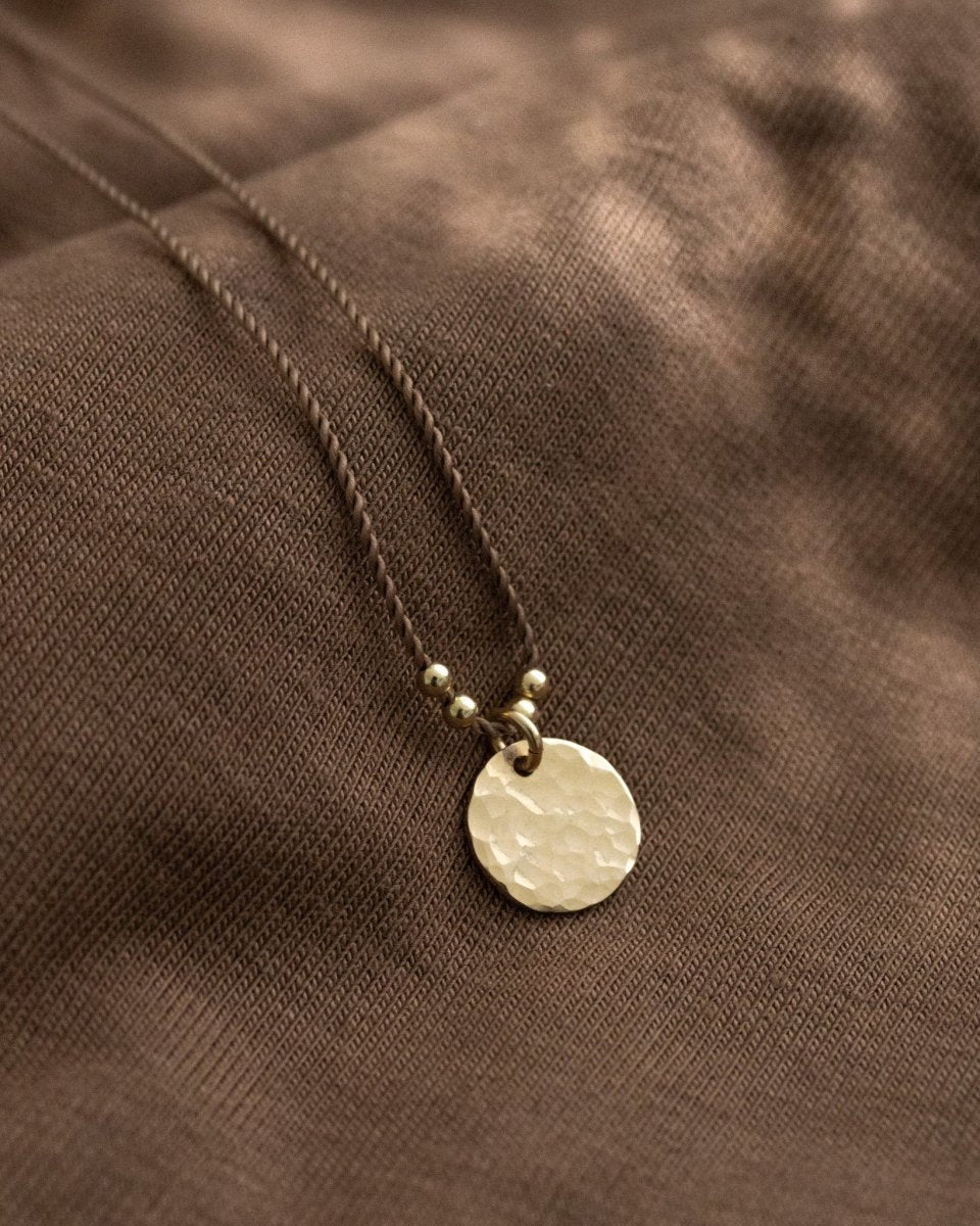 Eine braune Schnurkette mit einem gehämmerten, runden Plättchenanhänger in gold lehnt an braunem Stoff.