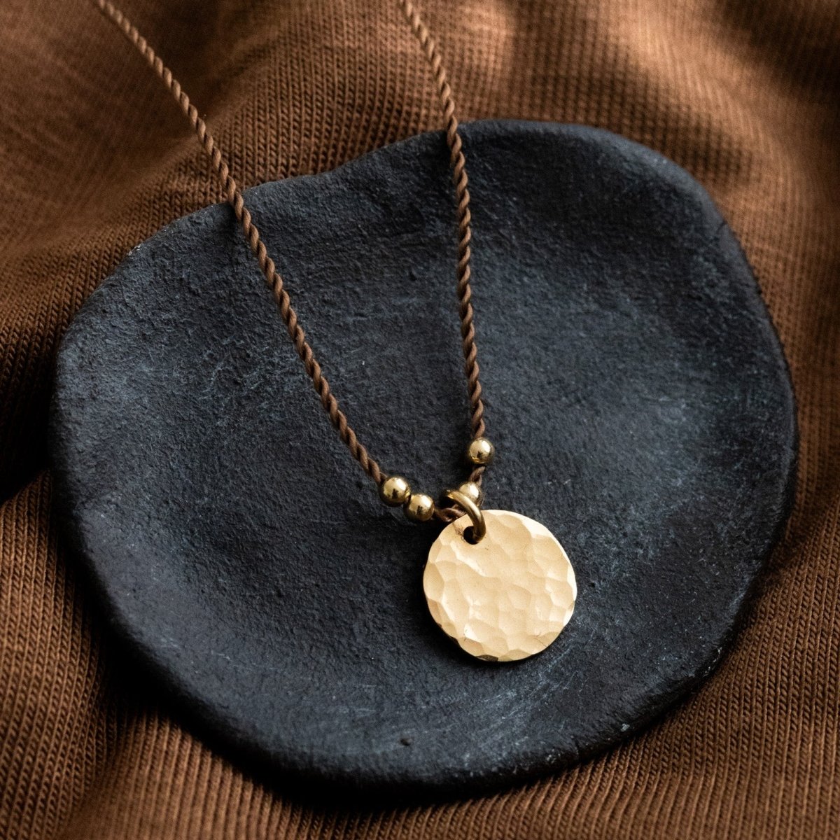 Eine braune Schnurkette mit einem gehämmerten, runden Plättchenanhänger in gold liegt auf einem schwarzen Steinschälchen.