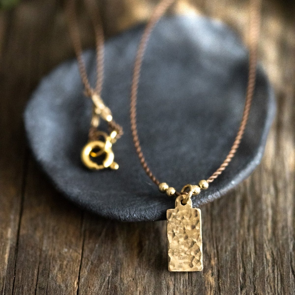 Eine feine, beige Kordelkette mit einem rechteckigen, gehämmerten Plättchenanhänger lehnt an einem dunkelgrauen Steinschälchen auf braunem Holzuntergrund.