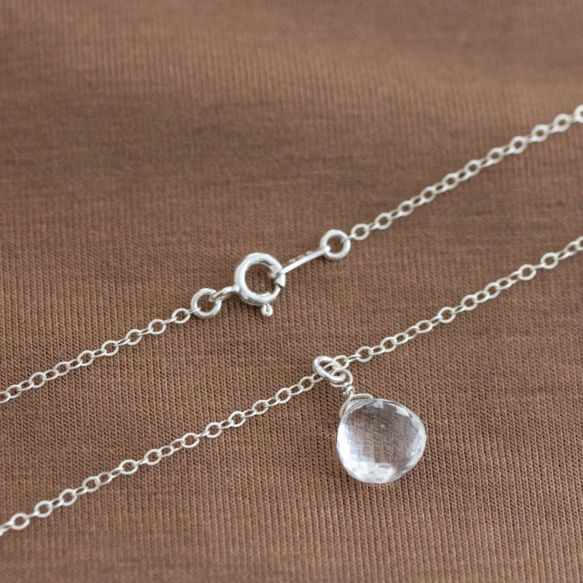 Eine feine Silberkette mit Bergkristall-Anhänger und Federring-Verschluss liegt auf einem braunen Stoff.
