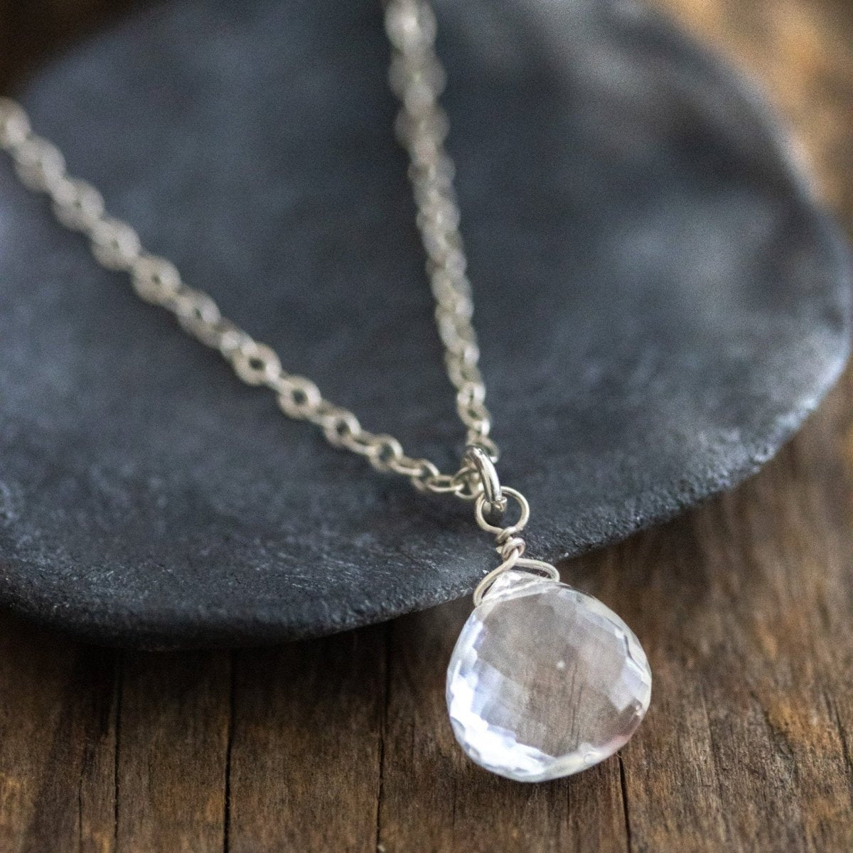 Eine dünne Silberkette mit einem Bergkristall-Anhänger in bauchiger Tropfenform lehnt an einem grauen Schälchen auf Holzuntergrund.
