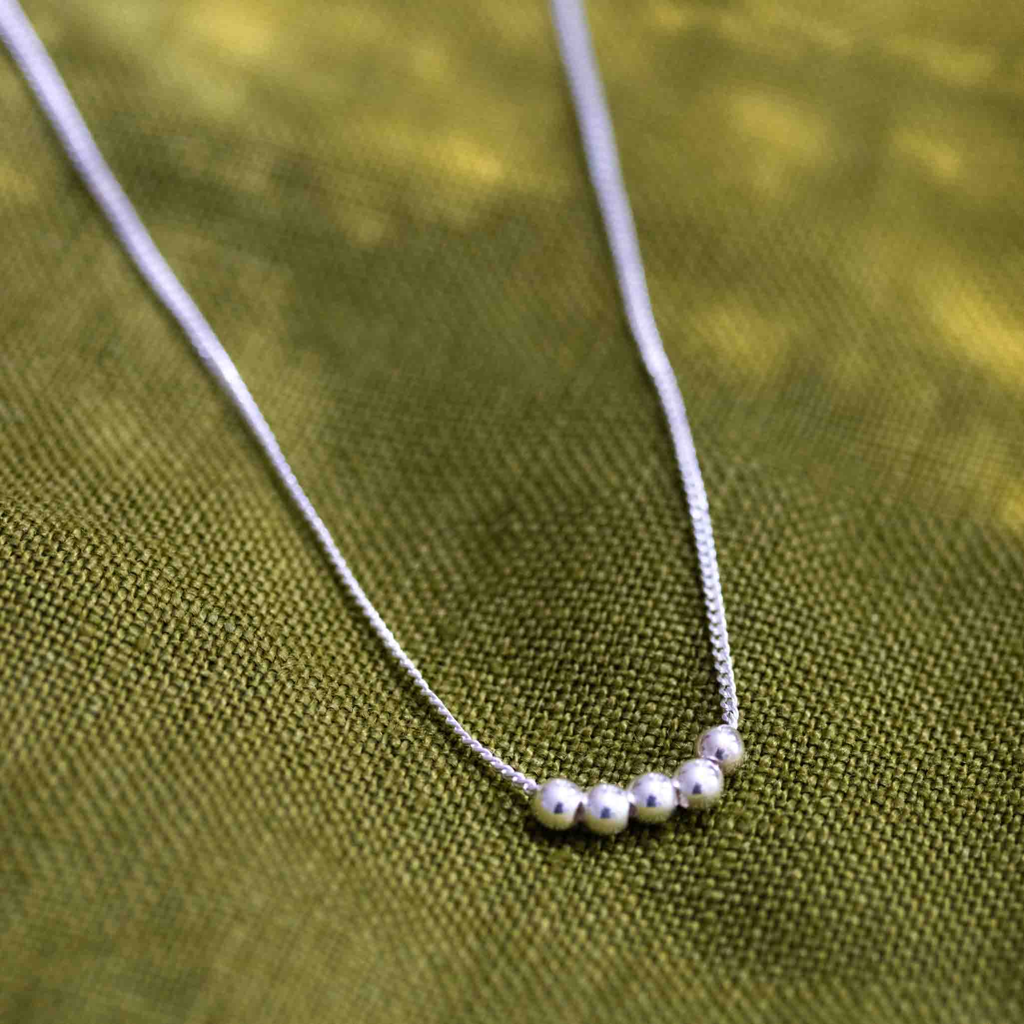 Eine feine Silberketten mit fünf kleinen Silberperlen liegt auf einem grünen Leinenstoff.