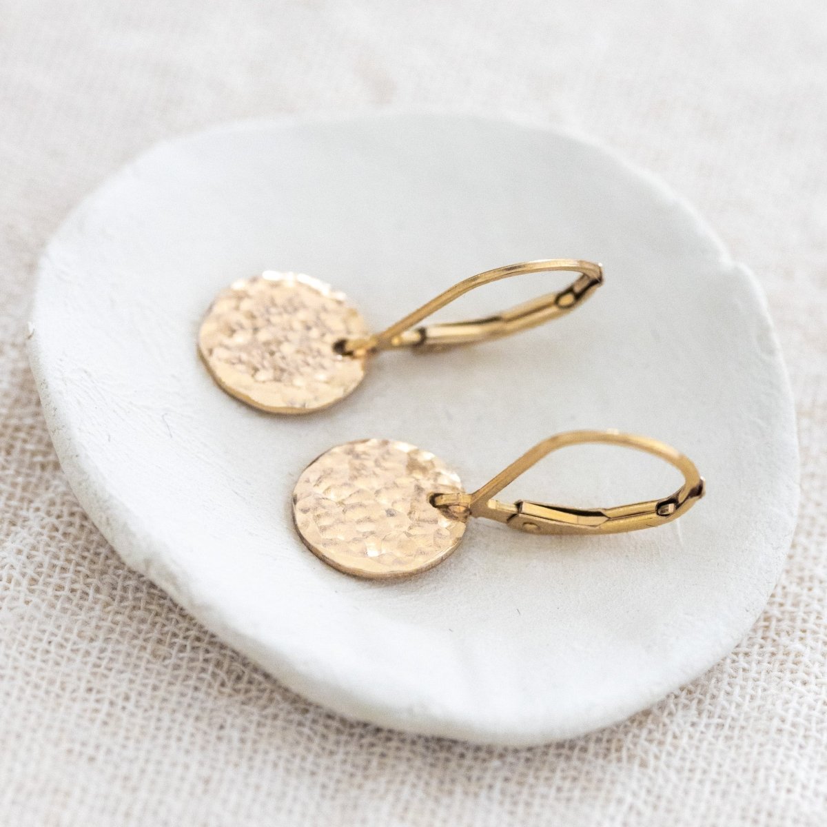 Ein Paar Gold-Ohrringe mit runden, gehämmerten Plättchen an Klappbrisur liegt auf einem weißen Schälchen.