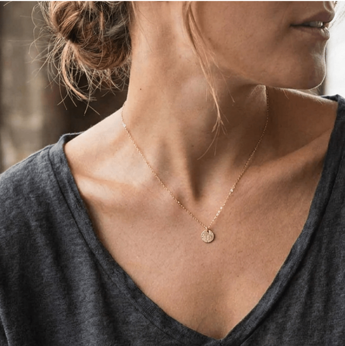 Eine feine und kurze Goldkette mit einem runden, gehämmerten Plättchen-Anhänger hängt vom Hals des Models herab.