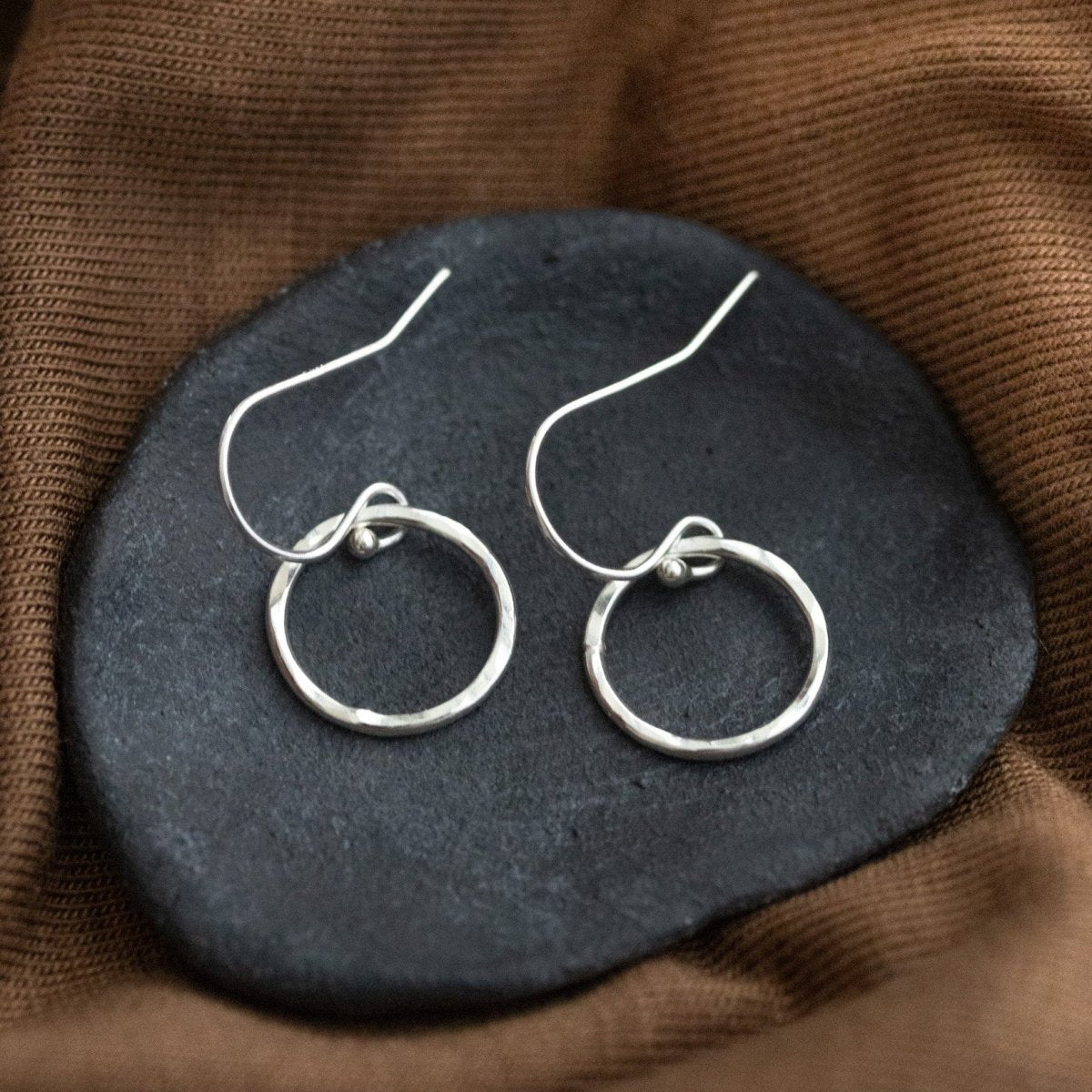 Ein Paar Silber-Ohrringe, bestehend aus jeweils einem gehämmerten Ring an Ohrhaken, liegt auf einem grauen Steinschälchen, das auf braunem Stoff liegt.