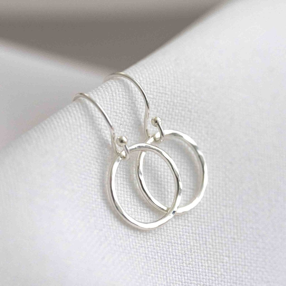 Ein Paar silberner Ohrringe bestehend aus einfachen Ohrhaken und gehämmerten Ringen auf weißem Hintergrund.
