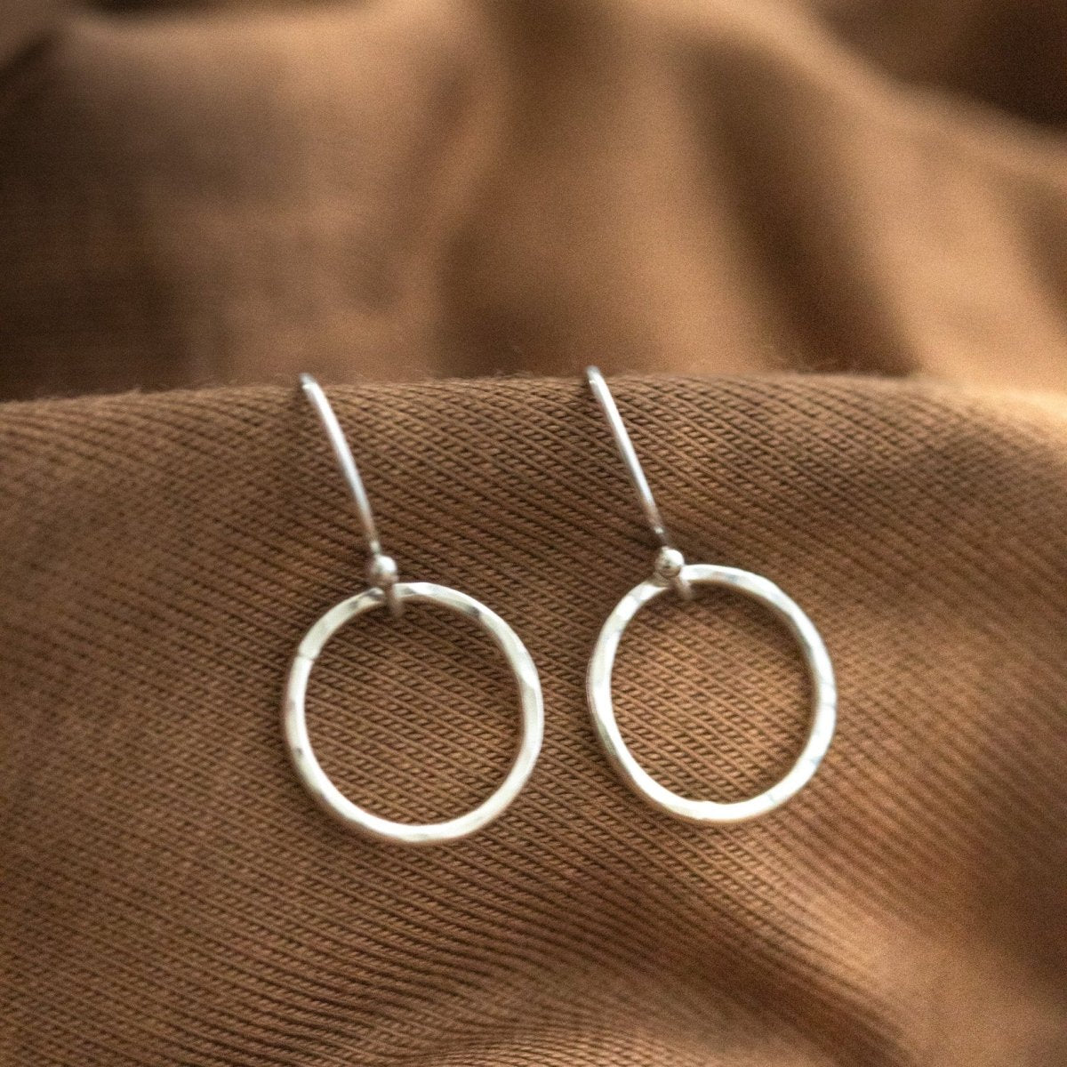 Ein Paar Silber-Ohrringe, bestehend aus jeweils einem gehämmerten Ring an Ohrhaken, hängt an gewelltem, braunen Stoff.