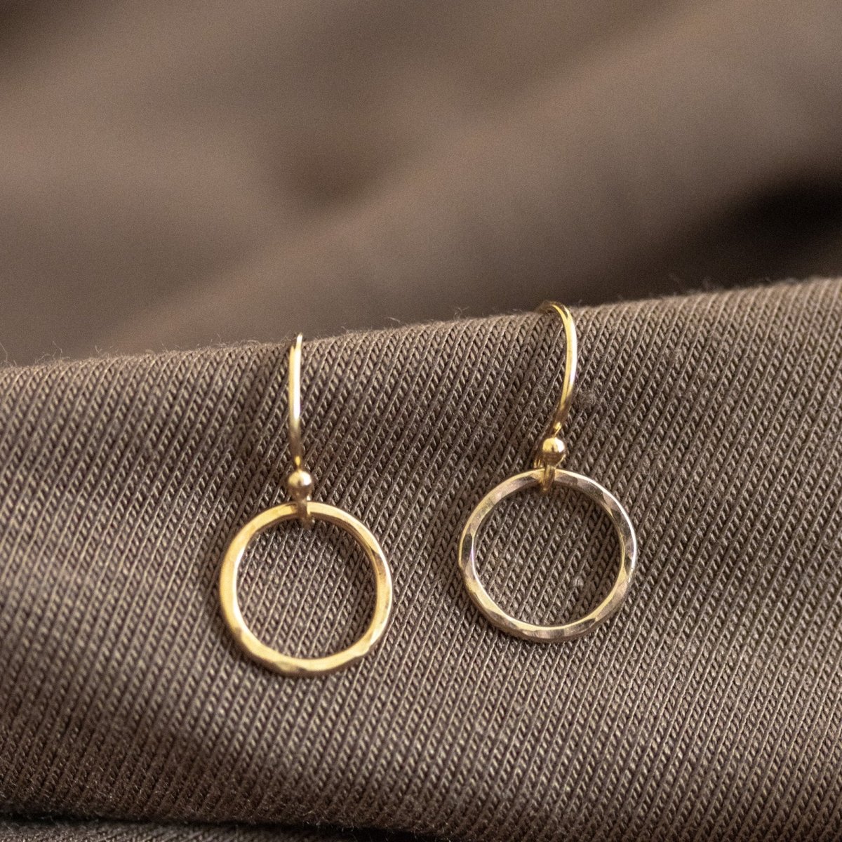 Ein Paar goldener Ohrringe, bestehend aus jeweils einem gehämmerten Ring an Ohrhaken, lehnt an gewelltem, braunen Stoff.