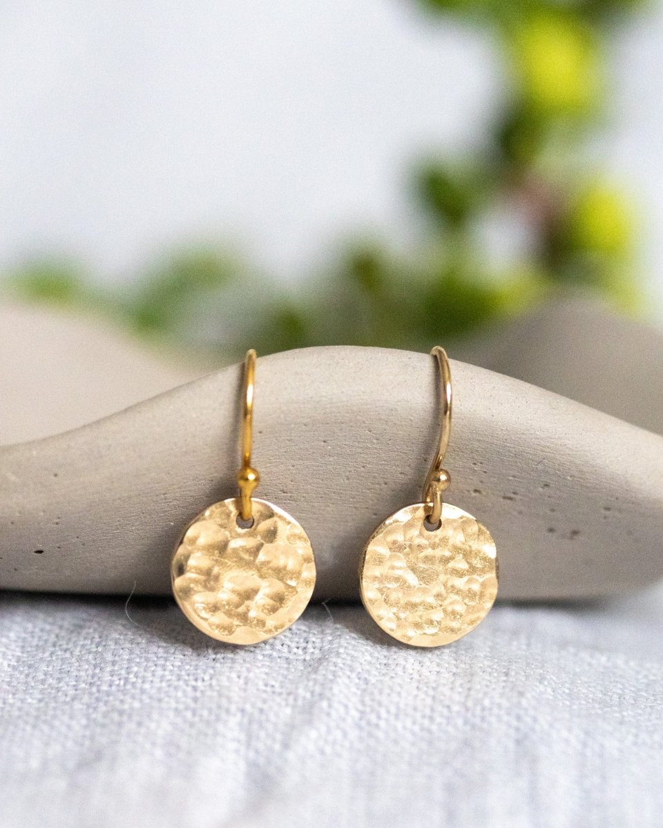 Ein Paar goldener Plättchen-Ohrringe in Hammerschlag-Optik hängen von einem kleinen, gewellten Schälchen herab. Verschwommenes Grün im Hintergrund.