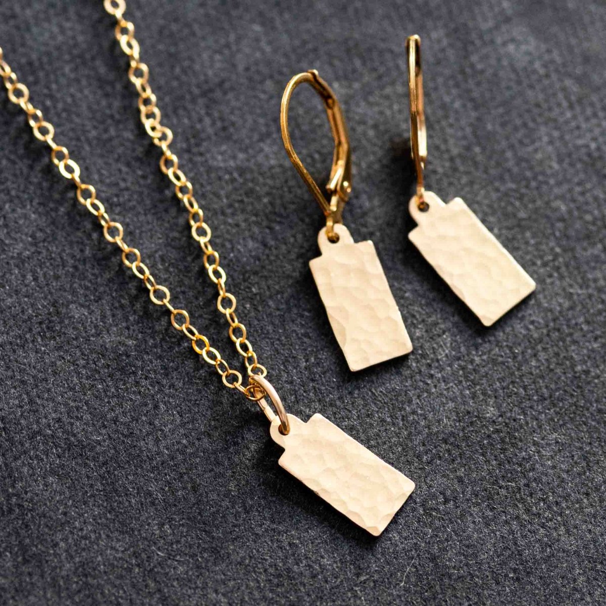 Ein goldenes Schmuckset auf grauem Hintergrund, bestehend aus einer Kette und Ohrringe mit rechteckigen Plättchen.