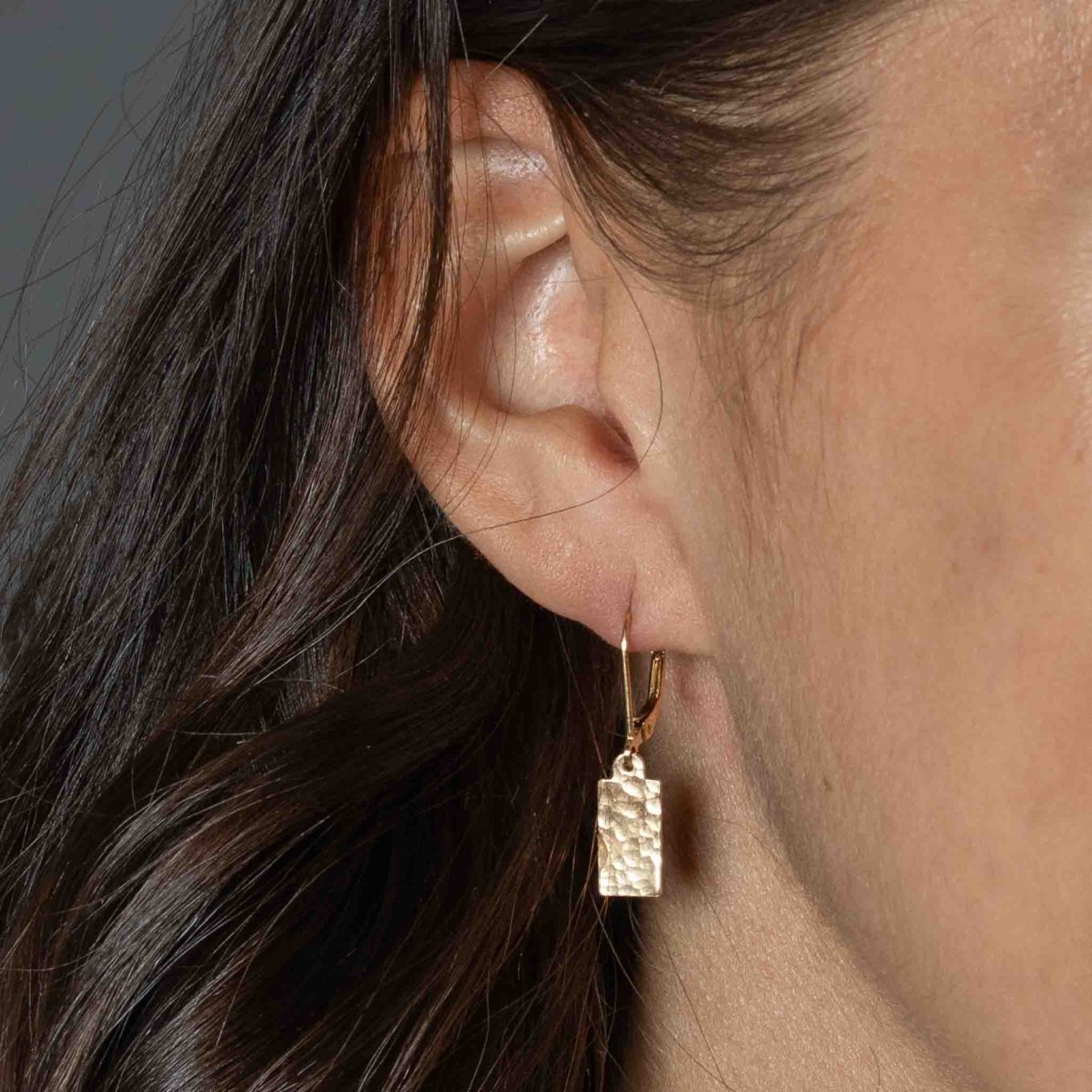 Am Ohr des Models hängt ein goldener Ohrring mit einem rechteckigem, gehämmerten Plättchen.