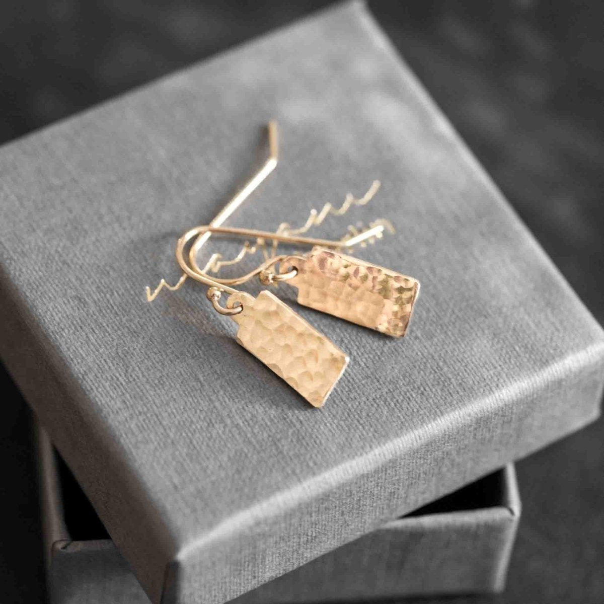 Auf einem grauen Schmucketui liegt ein Paar goldener Ohrringe mit gehämmerten, rechteckigen Plättchen-Anhängern.