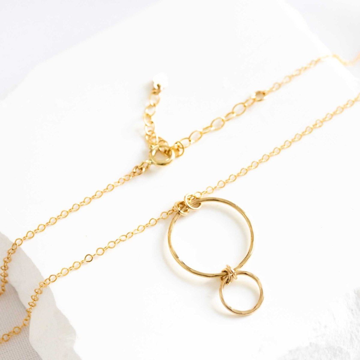 Eine feine Goldkette mit einem Anhänger, bestehend aus einem größeren Ring, an dem ein kleinerer Ring hängt, liegt auf weißem Hintergrund.