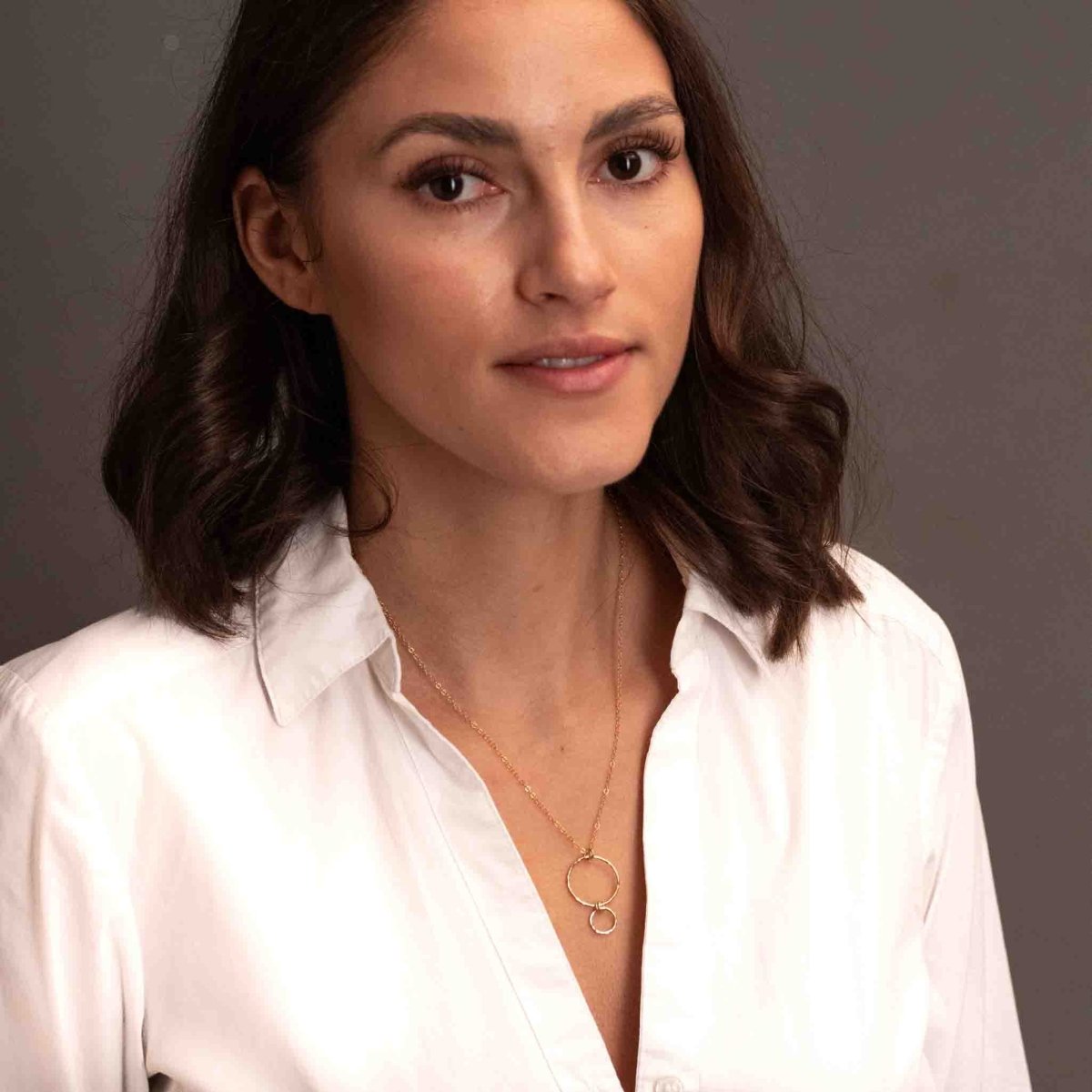 Eine feine Goldkette mit einem Anhänger, bestehend aus einem größeren Ring, an dem ein kleinerer Ring hängt, hängt vom Hals des Models.