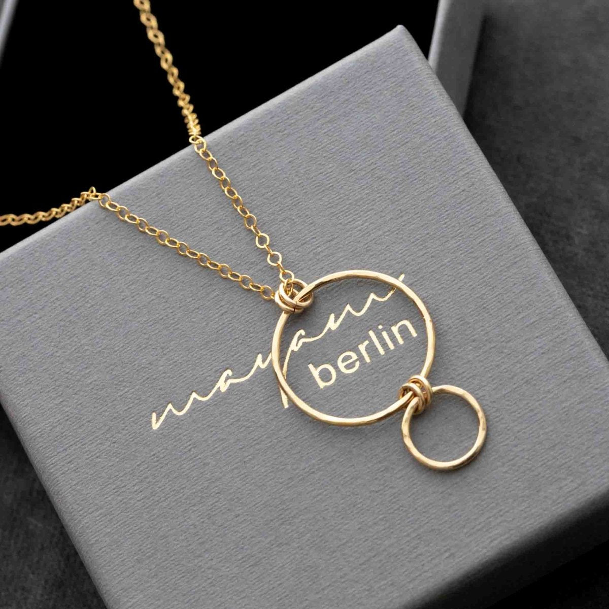 Eine feine Goldkette mit einem Anhänger, bestehend aus einem größeren Ring, liegt auf einem grauen Schmuck-Etui, in das der Name mayam berlin eingeprägt ist.