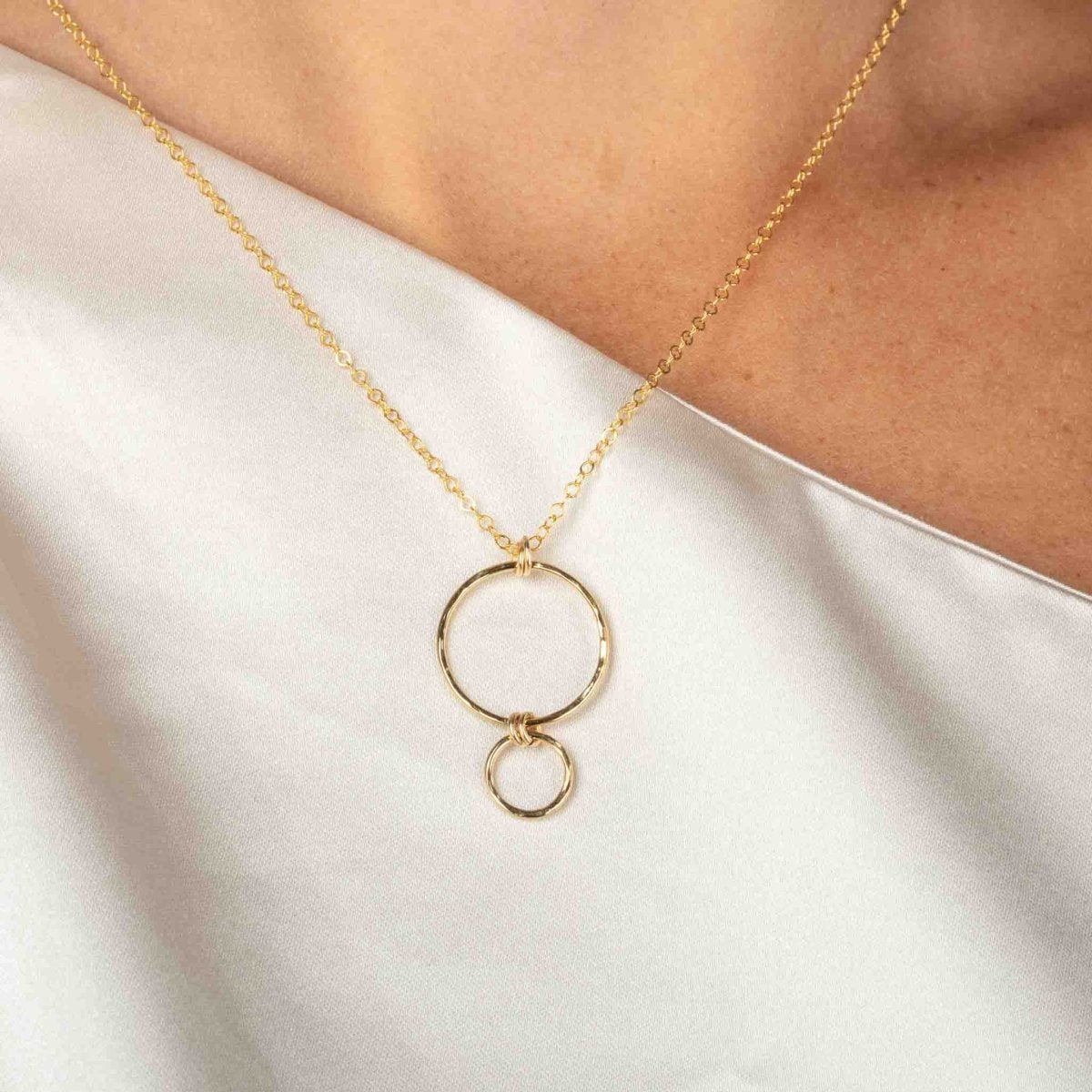 Eine feine Goldkette mit einem Anhänger, bestehend aus einem größeren Ring, an dem ein kleinerer Ring hängt, hängt vom Hals des Models.