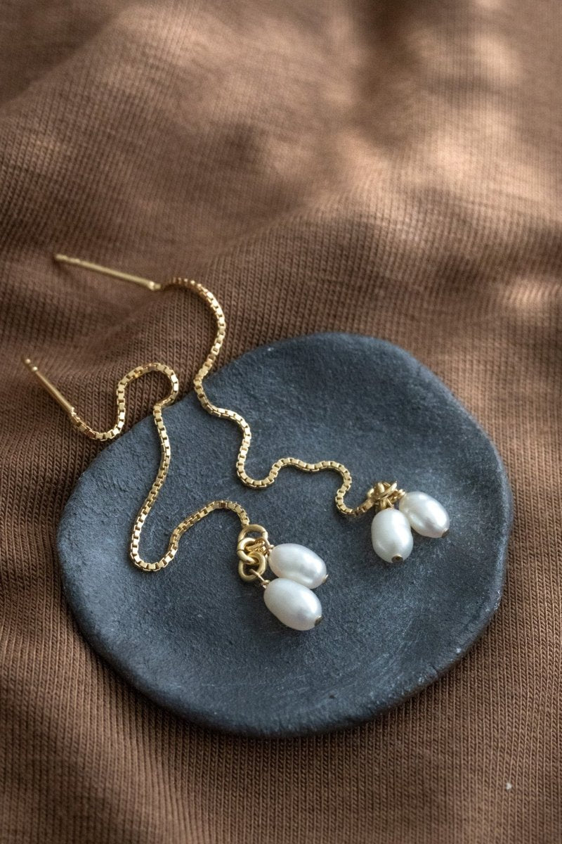 Ein Paar goldener Einfädler Ohrringe mit jeweils zwei kleinen Keshi-Perlen liegt auf einem Schälchen auf braunem Stoff.