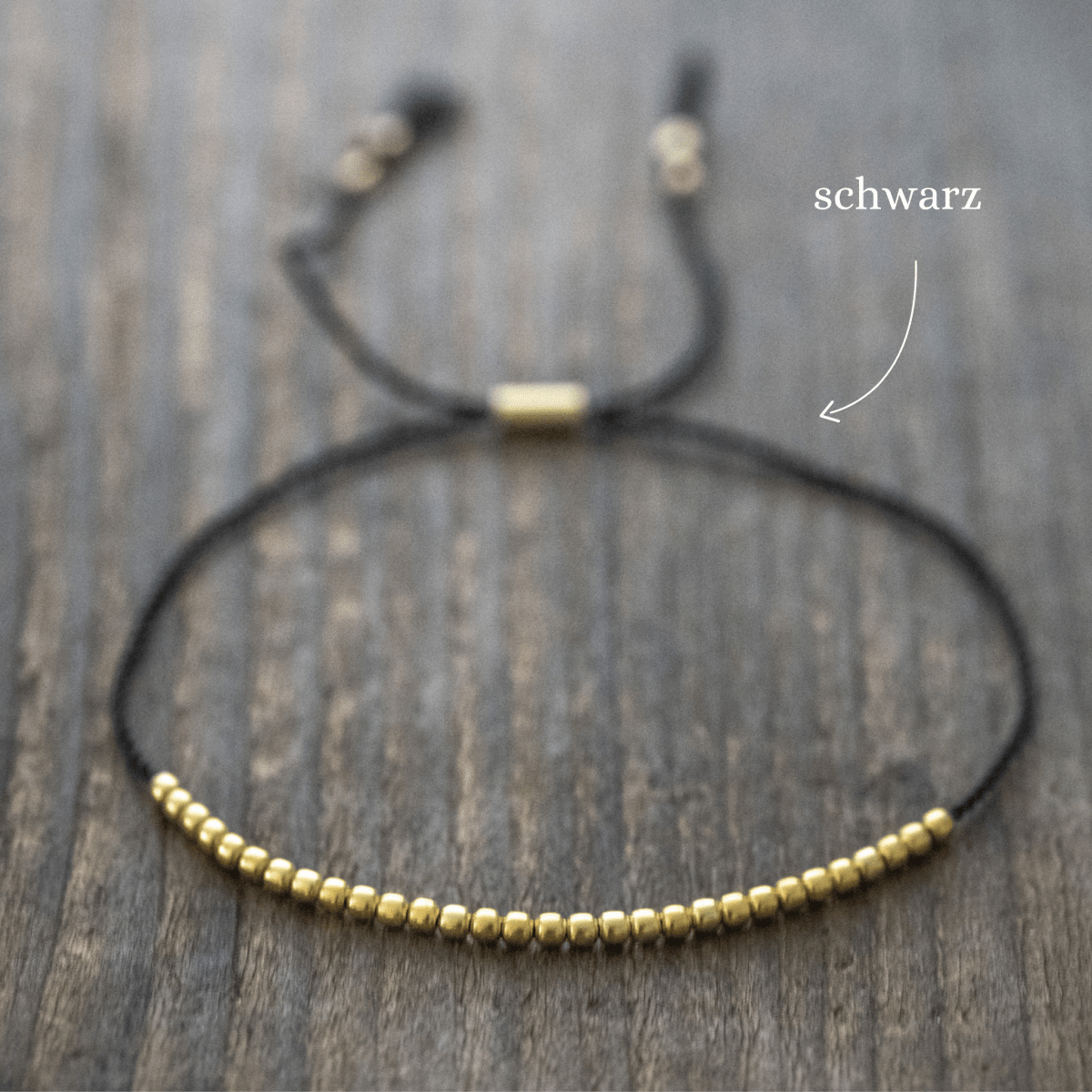 Ein dünnes, schwarzes Schnurarmband mit vielen goldenen Perlen und einem Schiebeverschluss liegt auf braunem Holz.