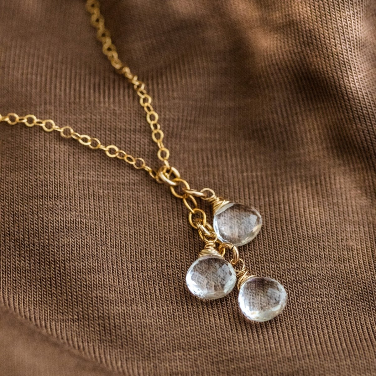 Eine feine Goldkette mit drei untereinander angebrachten, kleinen Bergkristalltropfen liegt auf braunem Stoff.
