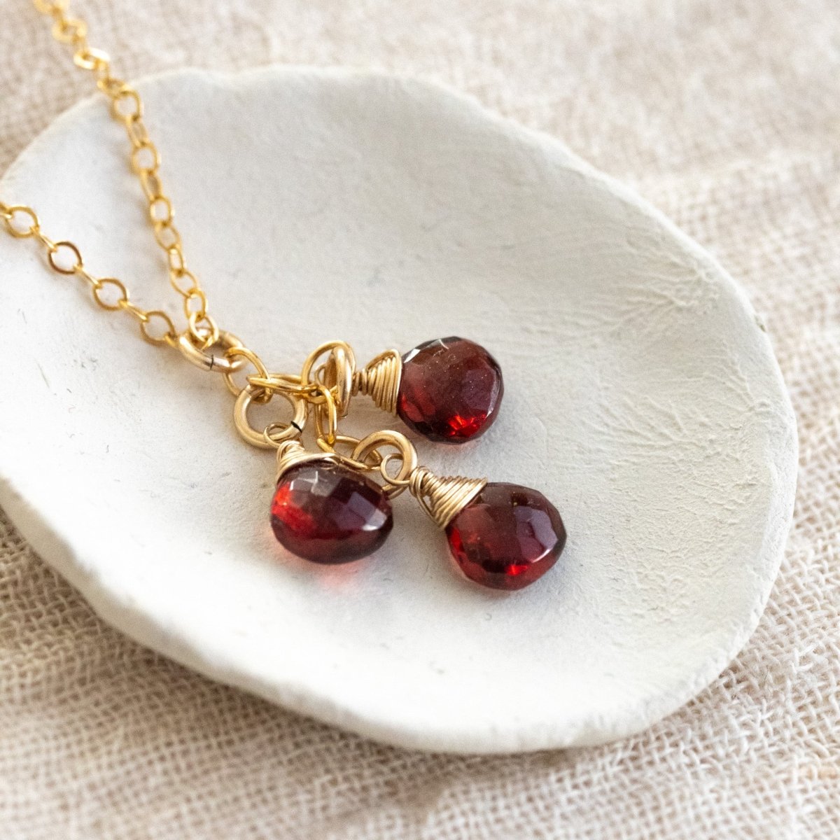 Eine feine Goldkette mit drei untereinander hängenden Granat Edelsteinen liegt auf einem weißen Schälchen.