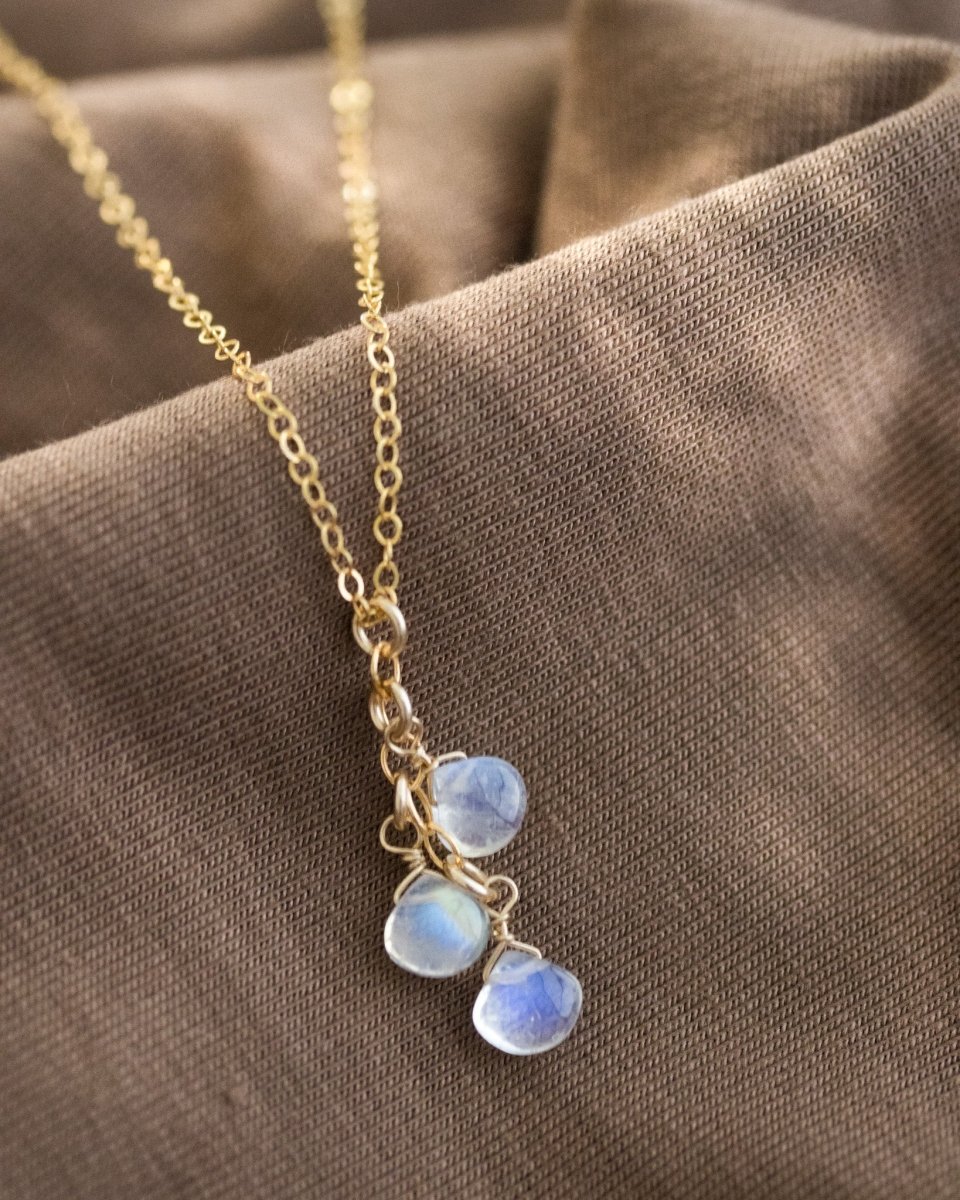 Eine feine Goldkette mit drei untereinander hängenden Mondsteintropfen lehnt an einem gewellten, braunen Stoff.
