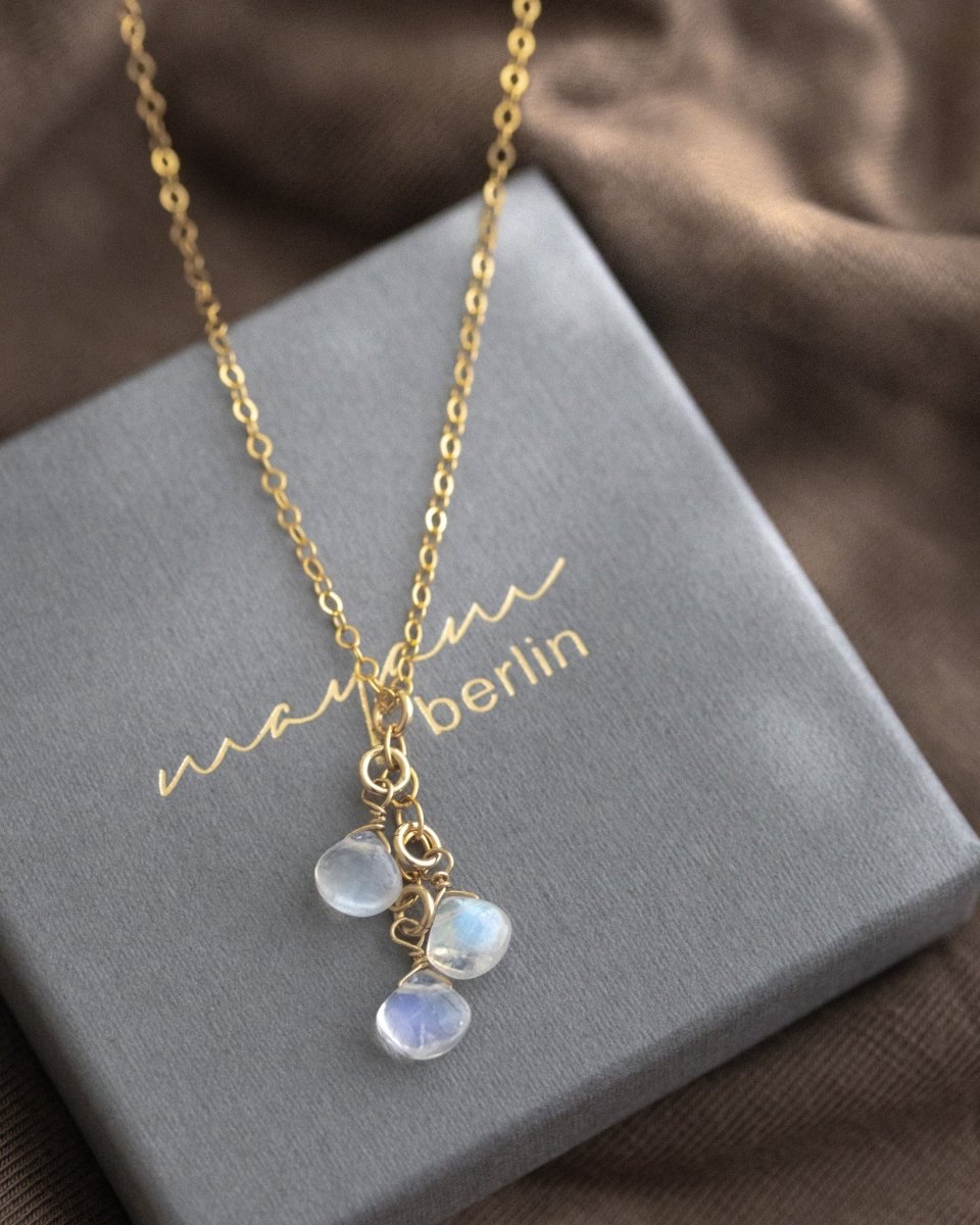 Eine feine Goldkette mit drei untereinander hängenden Mondsteintropfen liegt auf einem grauen Schmucketui.