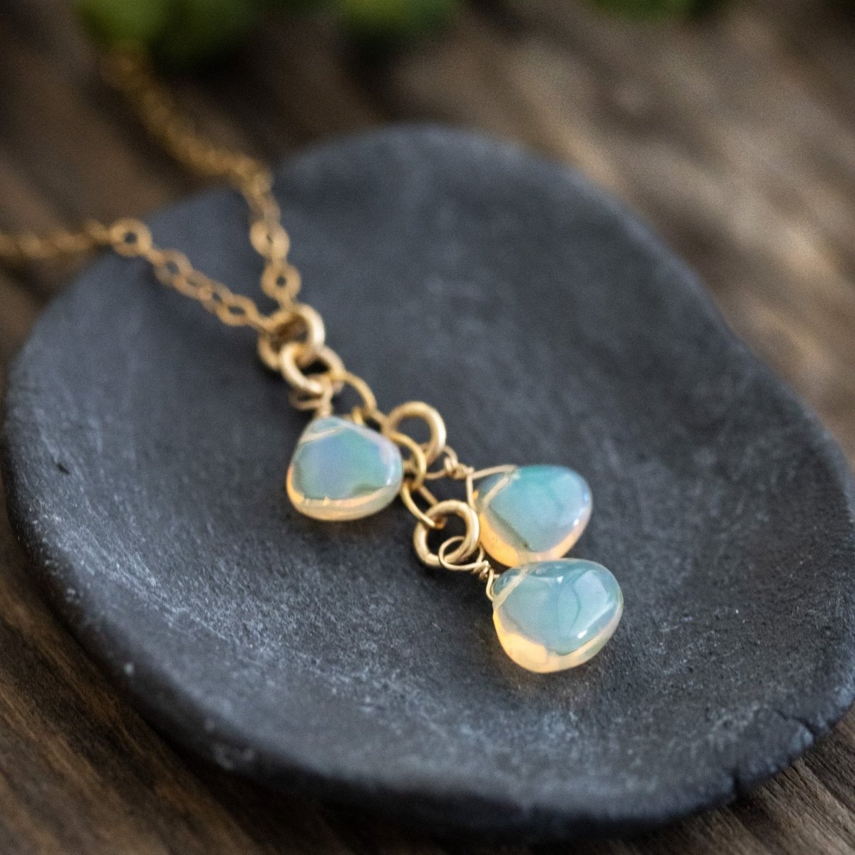 Eine feine Goldkette mit drei Opal-Anhänger liegt auf einem dunkelgrauen Schälchen, Holzuntergrund.