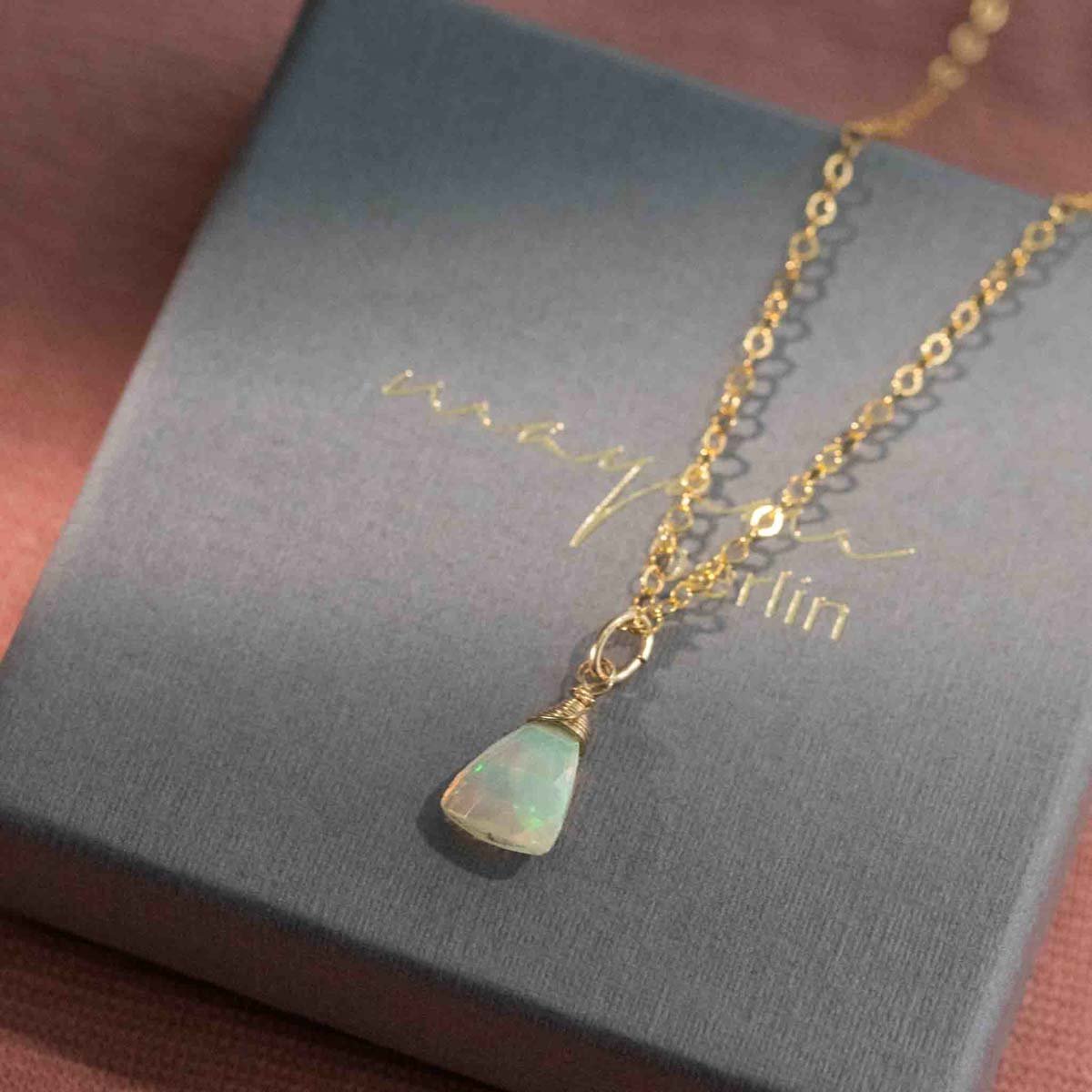 Eine zierliche Goldkette mit einem dreieckigen Welo Opal-Anhänger liegt auf einem grauen Schmucketui.