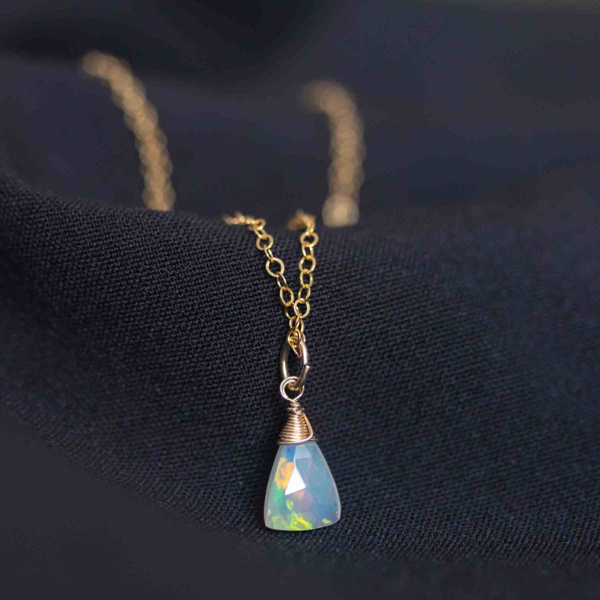 Eine zierliche Goldkette mit einem dreieckigen Welo Opal-Anhänger liegt auf schwarzem Hintergrund.