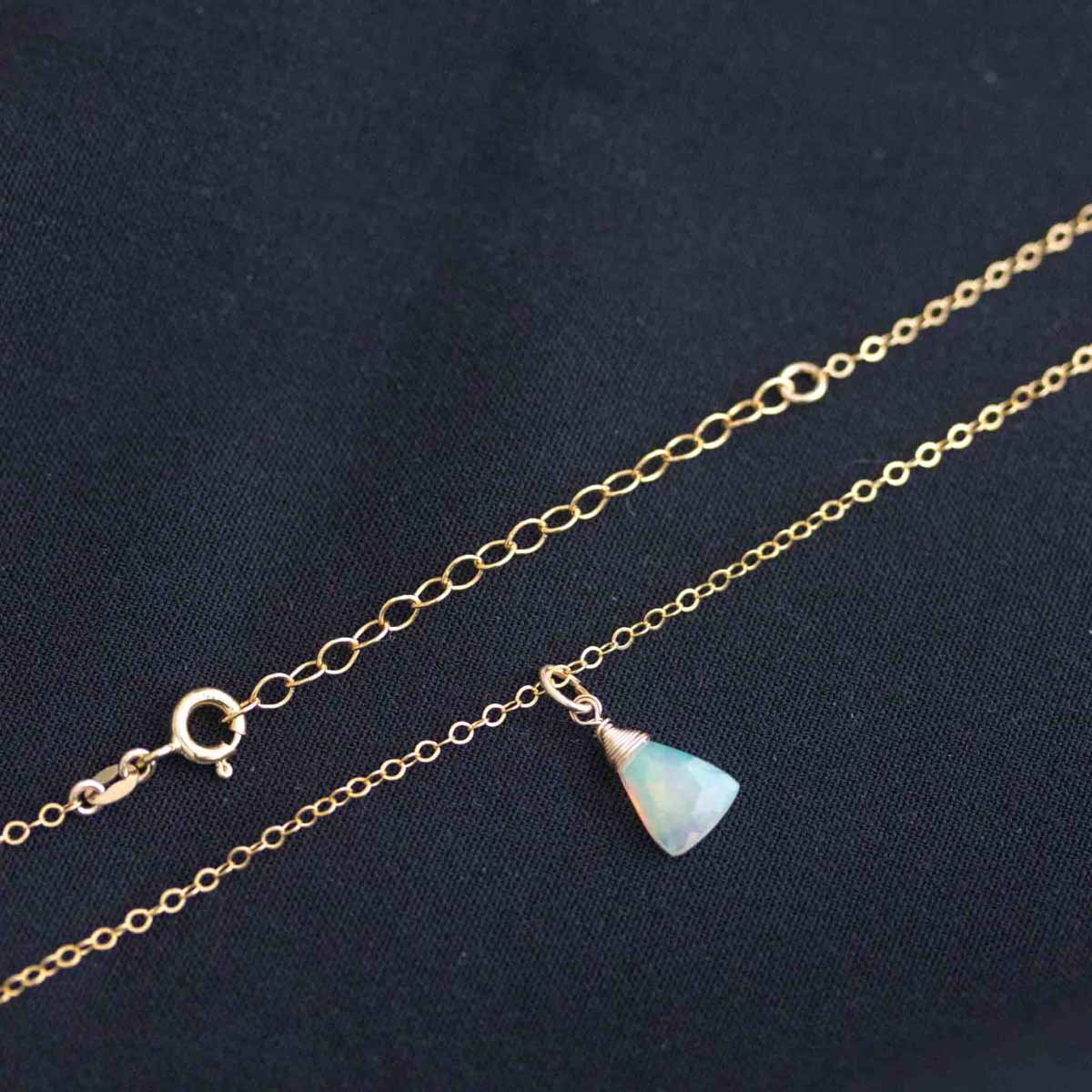 Eine zierliche Goldkette mit einem dreieckigen Welo Opal-Anhänger liegt auf schwarzem Hintergrund.