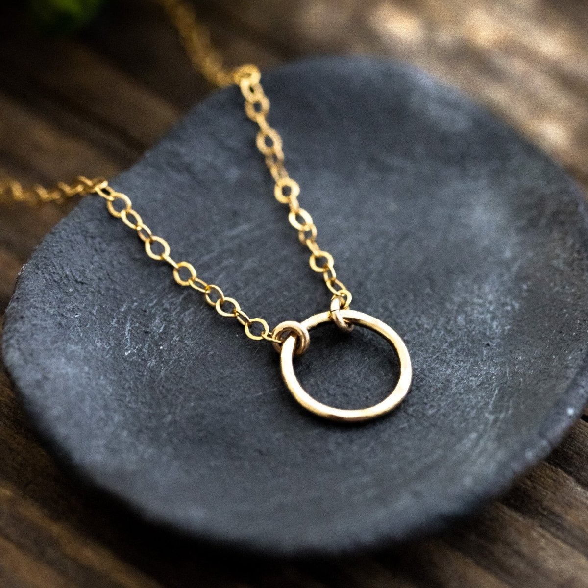 Eine feine, goldene Halskette mit einem Ring-Anhänger liegt auf einem grauen Steinschälchen, das auf Holzuntergrund steht.