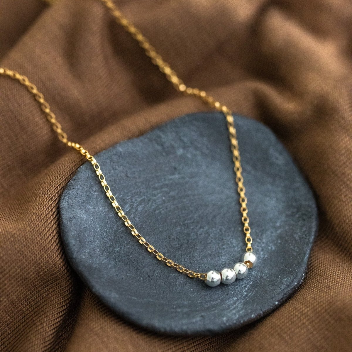 Eine Goldkette mit einer Reihe kleiner Silberperlen in der Mitte liegt auf einem schwarzen Schälchen.