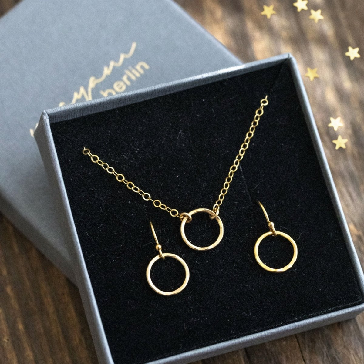 In einem offenen Schmucketui ist ein Set, bestehend aus feiner Goldkette und Ohrringen, eingehängt. An der Kette und an den Ohrringen hängt jeweils ein gehämmerter Ring. Auf dem Holzuntergrund liegt Sternchenkonfetti verstreut.
