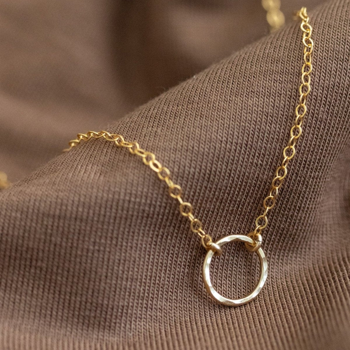 Eine feine Goldkette mit einem gehämmerten Ring im Zentrum lehnt an gewelltem, braunen Stoff.