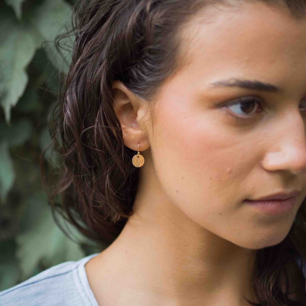 Am Ohr des Models hängt ein goldener Ohrring mit einem gehämmerten Plättchen.