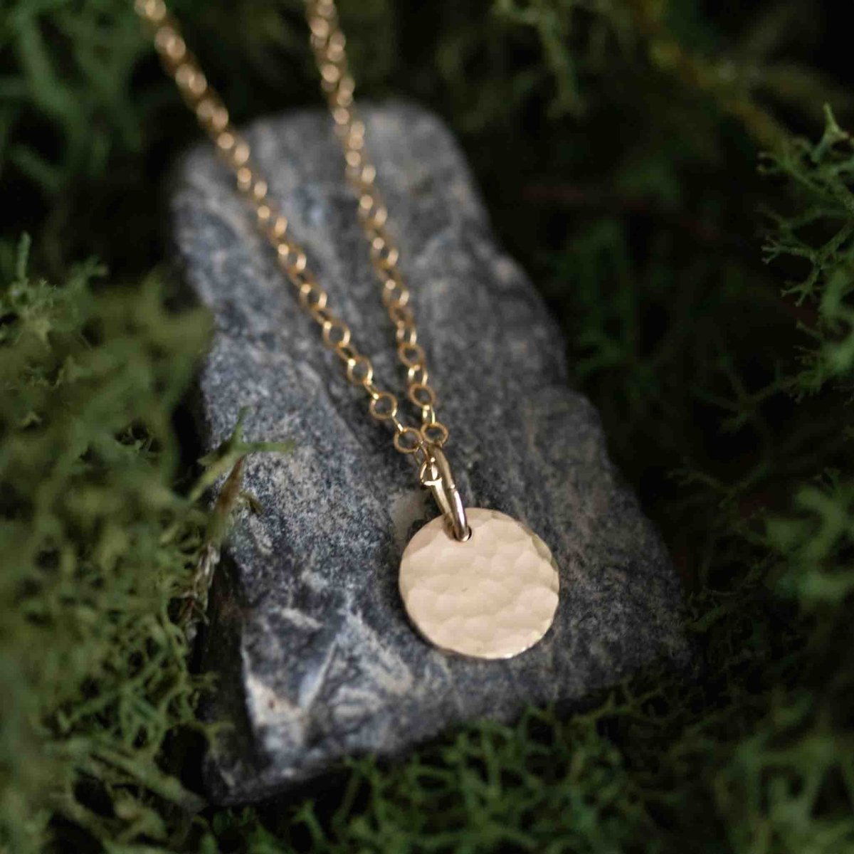 Eine feine Goldkette mit einem gehämmerten Plättchen-Anhänger liegt auf einem grauen Stein, der auf Moos liegt.