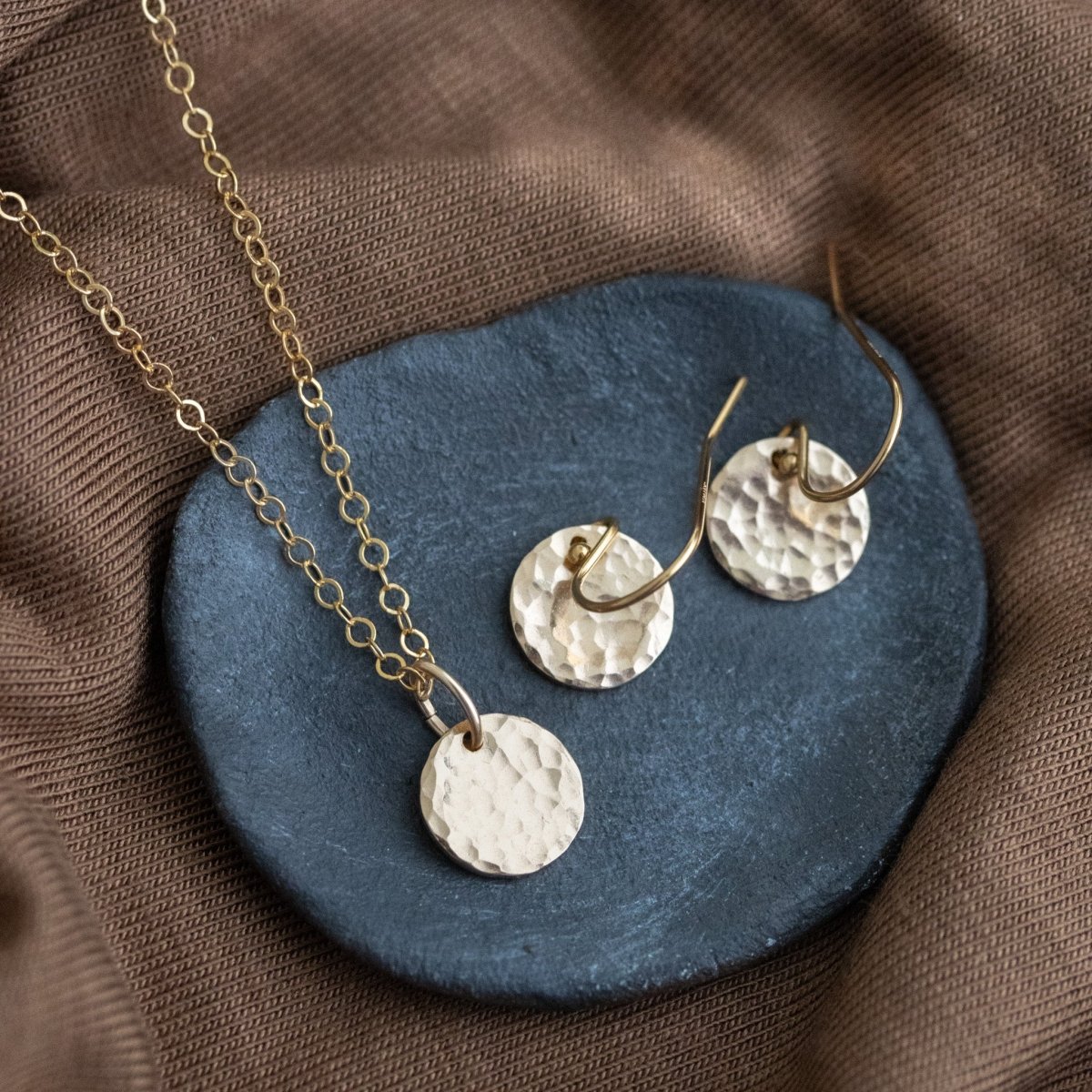 Ein goldenes Schmuckset, bestehend aus einer Kette und einem Paar Ohrringe mit gehämmerten Plättchen liegt auf einem grauen Steinschälchen, das auf braunem Stoff liegt.