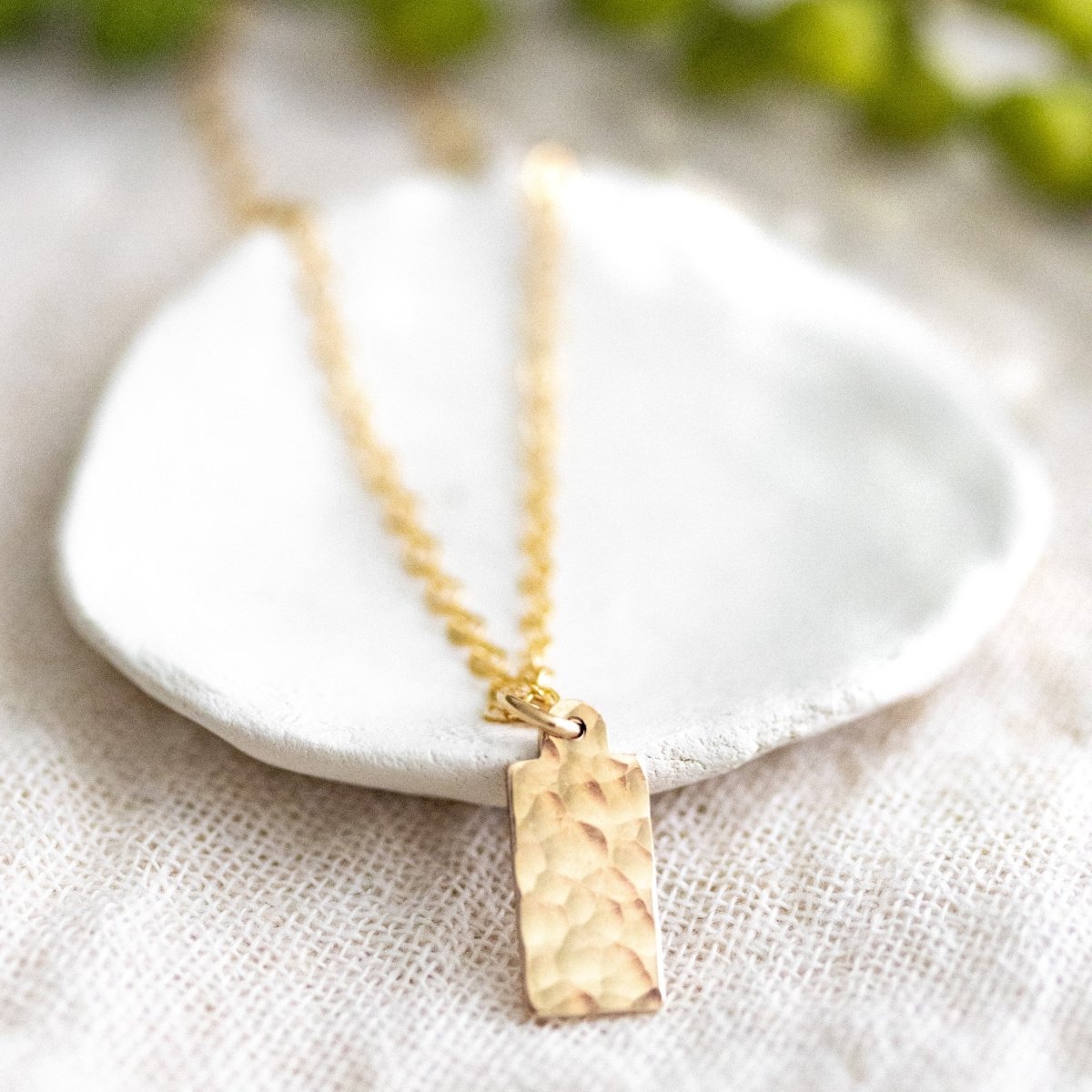 Gehämmertes, rechteckiges Goldplättchen an feiner Goldkette. Angelehnt an weißes Schälchen.