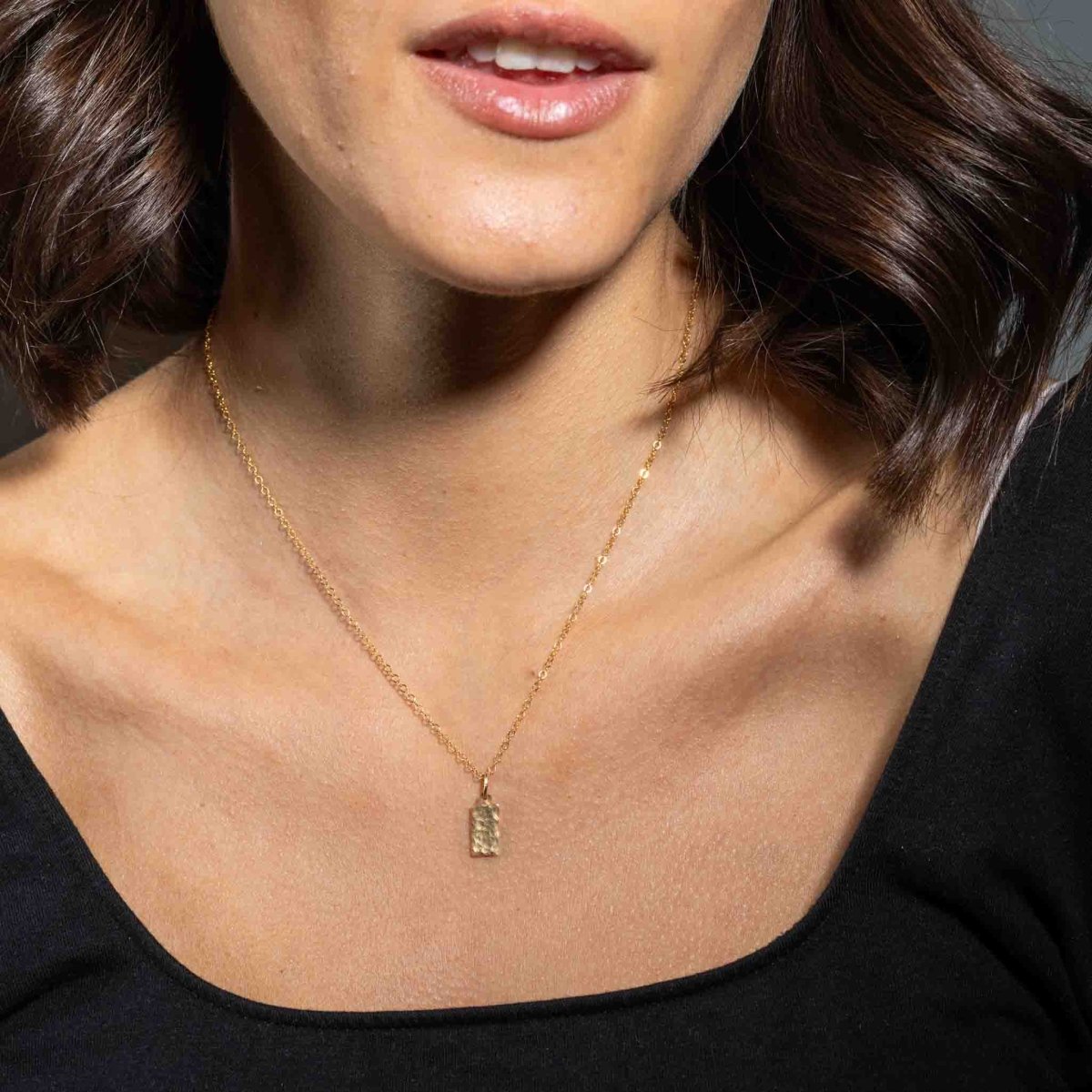 Eine feine und kurze Goldkette mit einem rechteckigen, gehämmerten Plättchen-Anhänger hängt vom Hals des Models herab.