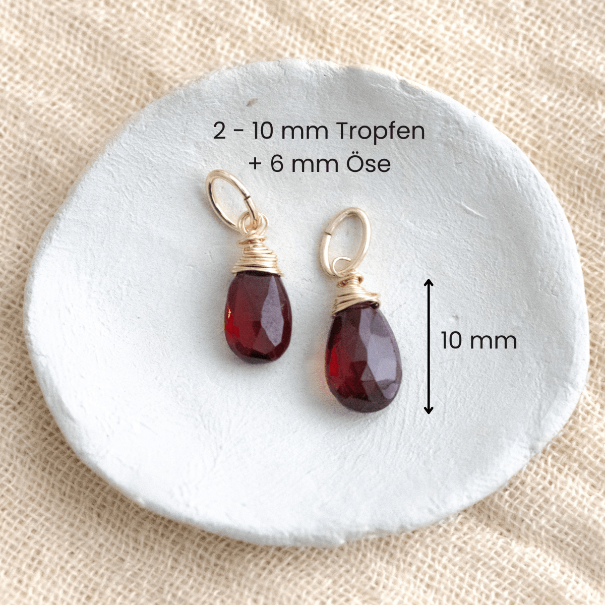 Zwei Granat-Anhänger an goldener Öse liegen in einem weißen Schälchen auf hellem Stoff, mit Maßangaben.