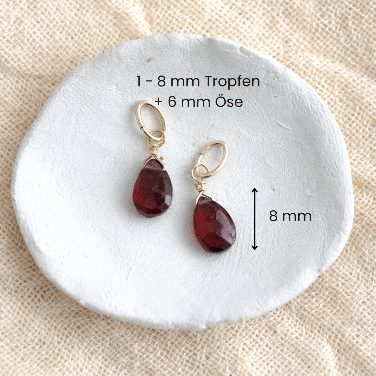 Zwei Granat-Anhänger an goldener Öse liegen in einem weißen Schälchen auf hellem Stoff, mit Maßangaben.