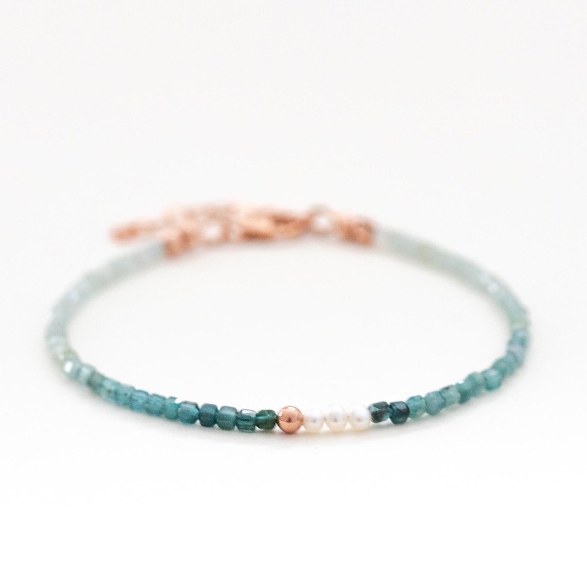 Ein dünnes Armband, bestehend aus kleinen Grandidierit-Perlen mit Farbverlauf und drei Süßwasserperlen in der Mitte liegt auf weißem Hintergrund.