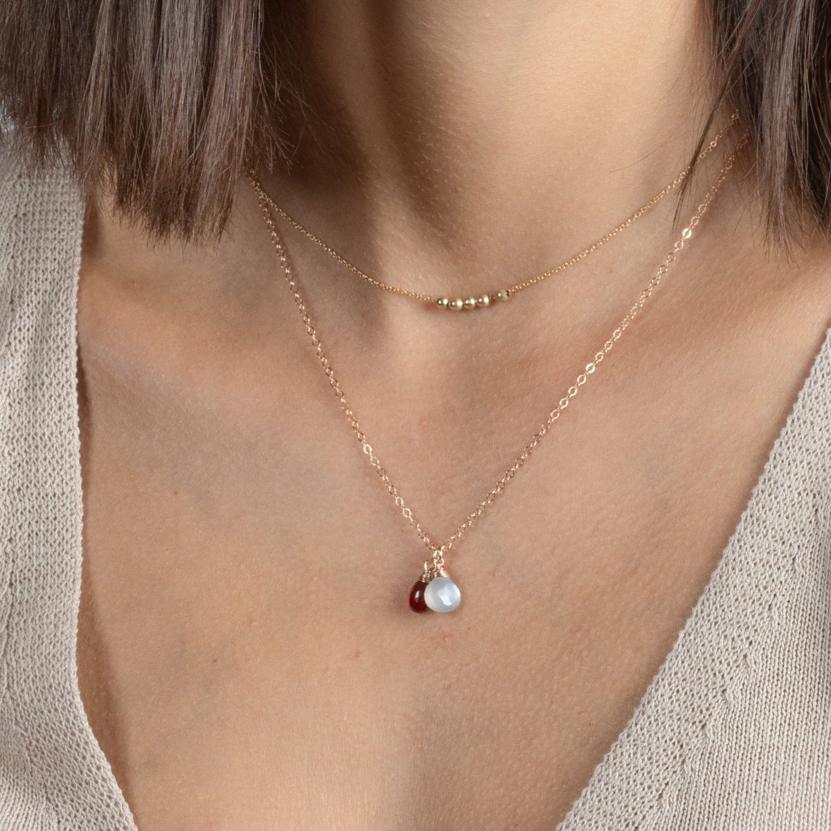 Am Hals des Models hängt eine kürzere Rosegold-Kette mit fünf Rosegold-Perlen und eine längere Roségold-Kette mit zwei Edelstein-Anhängern, einem grauen Mondstein und einem dunkelroten Granat.