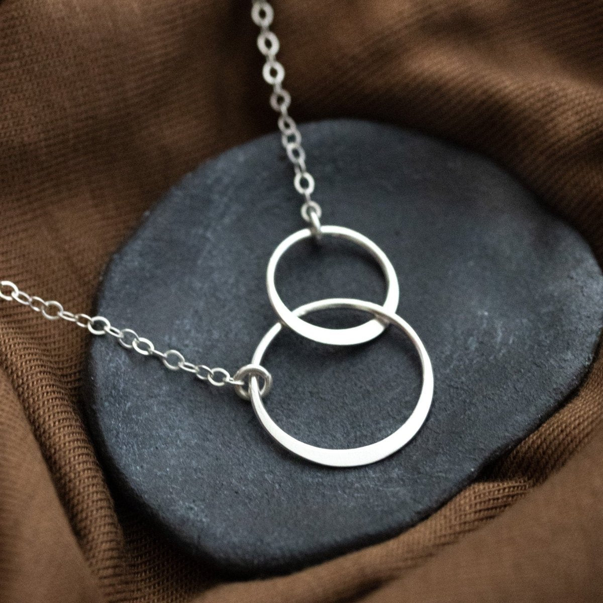 Eine zierliche 925 Silberkette mit zwei miteinander verschlungenen und unterschiedlich großen Ringen in der Mitte lehnt an einem grauen Schälchen auf gewelltem, braunen Stoff.